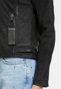 Schwarze Lederjacke mit seitlichem Reißverschluss und einem Markenetikett. Die Ärmel sind genäht und die Jacke wird mit hellblauen, mit Nieten verzierten Jeans kombiniert.