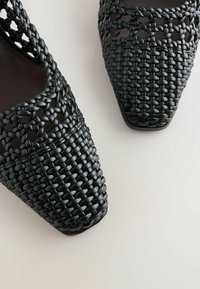 Chaussures sling-back en cuir tressé noir avec bouts carrés sur un fond clair, montrant en gros plan le motif tressé détaillé.