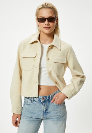 Beige taillierte Jacke mit weicher Textur, ausgestattet mit zwei Fronttaschen, einem Kragen und goldenen Knöpfen. Getragen über einem weißen Oberteil und blauen Jeans.