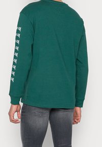 Vihreä pitkähihainen T-paita, joka on valmistettu puuvillasta. Oikeassa hihassa on valkoinen tekstikuvio. Rentoa istuvuutta, ribbaatut hihat ja pyöreä pääntie.