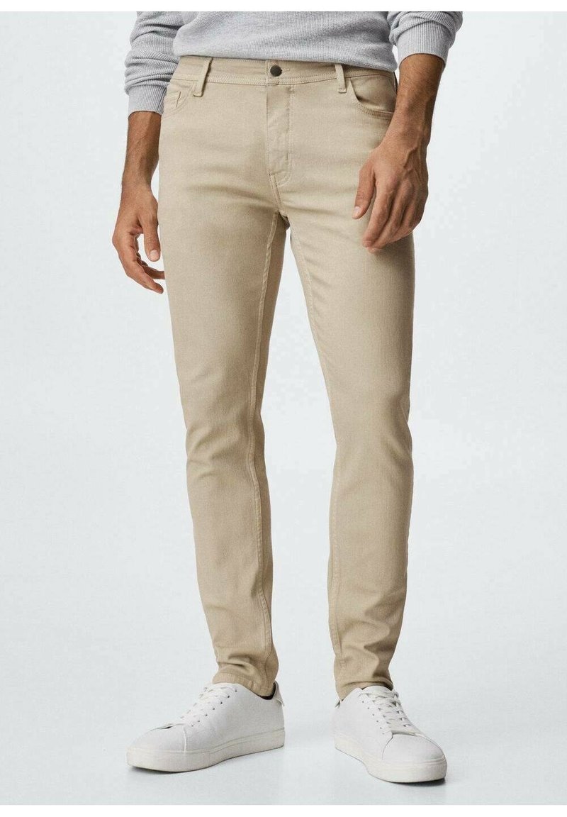 Homme portant un pantalon beige slim et des baskets blanches, debout devant un fond clair uni.