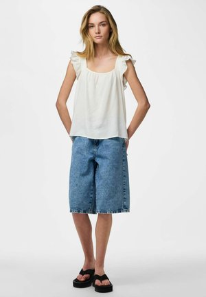 Giovane donna in piedi che indossa un top bianco senza maniche con volant, pantaloncini in denim al ginocchio e sandali neri con zeppa su sfondo semplice.