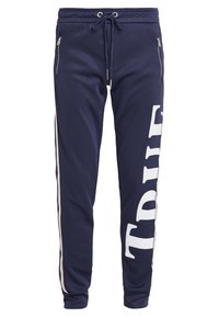 Mörkblå joggers tillverkade av mjukt tyg, med dragsko i midjan, sidofickor med dragkedja och en stor vit texttryck på vänster ben.