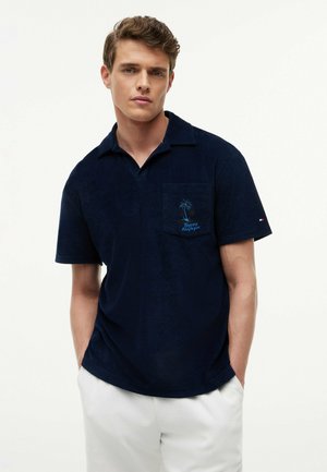 Młody mężczyzna w granatowej koszulce polo z haftem palm na kieszeni i białych spodniach, stojący z rękami w kieszeniach.