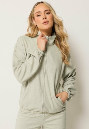 Frau mit langen blonden Haaren, die eine hellgraue, mit Reißverschluss versehene Jacke mit elastischen Bündchen und passende Hosen trägt, posiert mit einer Hand in der Tasche.