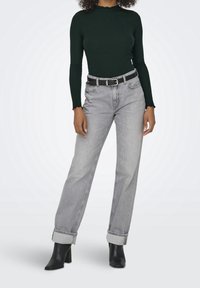 Ciemnozielona, prążkowana bluzka z długim rękawem i falbaniastymi brzegami, zestawiona z jasnoszarymi, szerokimi spodniami dżinsowymi i czarnymi botkami na obcasie. Szczegół z czarnym paskiem.