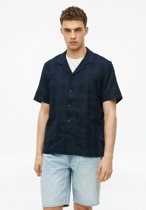 Jeune homme portant une chemise à manches courtes bleu foncé boutonnée sur un T-shirt blanc avec un short en jean bleu clair, debout devant un fond blanc.