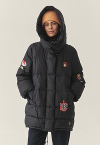 Juodas pūkinis džemperis su aukštu apykakle ir dideliu gobtuvu, dekoruotas ryškiais siuvinėtais raišteliais ant rankovių ir priekinės dalies. Tekstūruota medžiaga.