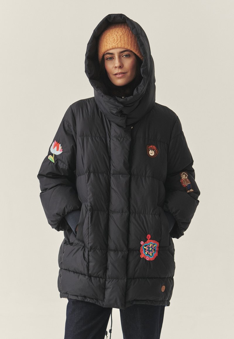 Juodas pūkinis džemperis su aukštu apykakle ir dideliu gobtuvu, dekoruotas ryškiais siuvinėtais raišteliais ant rankovių ir priekinės dalies. Tekstūruota medžiaga.