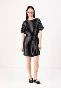 Vestido listrado em preto e branco com mangas curtas, faixa na cintura e um corte descontraído. O modelo usa sapatilhas pretas com detalhes decorativos.