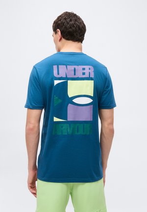 Mężczyzna w niebieskiej koszulce Under Armour z kolorowym logo blokowym na plecach i jasnooliwkowymi szortami, stojący na jednolitym białym tle.