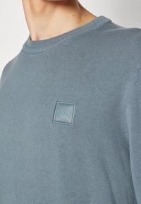 Gros plan sur une personne portant un pull en tricot bleu clair avec un petit patch logo BOSS brodé sur la poitrine.