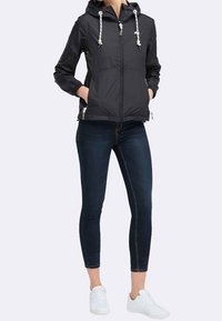 Donna che indossa una giacca nera con cappuccio e lacci bianchi, jeans skinny scuri, sneakers bianche, mani in tasca, in piedi contro uno sfondo neutro.