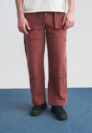 Άνδρας που φοράει ξεθωριασμένο κόκκινο παντελόνι τύπου cargo με μπροστινές και πλαϊνές τσέπες, μαύρα αθλητικά παπούτσια, στέκεται σε μπλε χαλί μπροστά σε λευκό τοίχο.