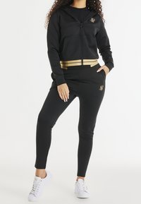 Zwarte hoodie met rits en gouden accenten en logo, gecombineerd met zwarte, aansluitende broek. Heeft een gladde textuur en een sportief ontwerp met zakken.