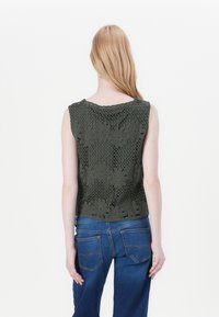 Olivengrønn heklet topp med blomstermønstre og bølgete kanter, med en avslappet passform. Kombinert med blå denimjeans, sett fra baksiden.