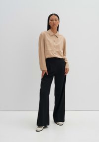 Beige knoopshirt met lange mouwen, zwarte hooggepaste broek en crèmekleurige schoenen. Het shirt heeft een kraag en voorzakken.