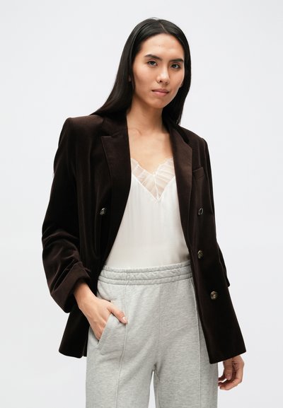 Hnedé zamatové dvojradové sako so zlatými gombíkmi, preložené cez bielu blúzku s čipkovaným lemom, v kombinácii so sivými teplákmi.