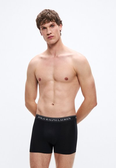 Fekete boxeralsók logóval ellátott derékpánttal. Az anyag simának tűnik, a fazon testhezálló, hossza combközépig ér. Minimalista stílus.