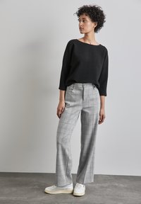 Svart texturerad stickad tröja med vid halsringning, matchad med grå rutiga högmidjade byxor och vita sneakers. Modell som står i profil.