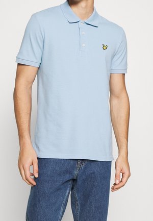 Mann trägt ein hellblaues kurzärmeliges Poloshirt mit Kragen und drei Knöpfen, kombiniert mit blauen Jeans, steht vor einem einfarbigen Hintergrund.