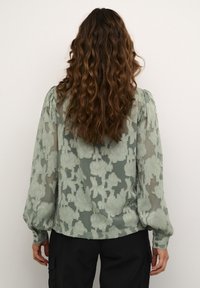 Sheer green blouse featuring textured floral embroidery, puff sleeves, and buttoned cuffs, paired with black pants, viewed from the back.