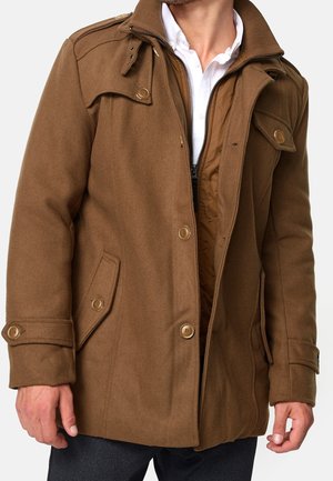 Homme portant un manteau en laine marron boutonné par-dessus une veste matelassée zippée et une chemise blanche à col, les mains détendues le long du corps.