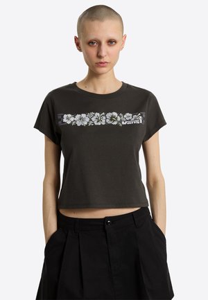 Kvinde med barberet hoved iført sort cropped t-shirt med hvidt blomsterdesign og sorte højttaljede bukser med hænderne i lommerne.