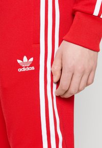 Röd adidas träningsoverall med vit tre-stripesdesign längs sidan. Har strukturerad tyg och en liten vit adidas-logga på benet.