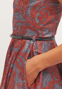 Gros plan d’une main dans la poche d’une robe grise avec un motif floral orange et une fine ceinture noire à la taille.