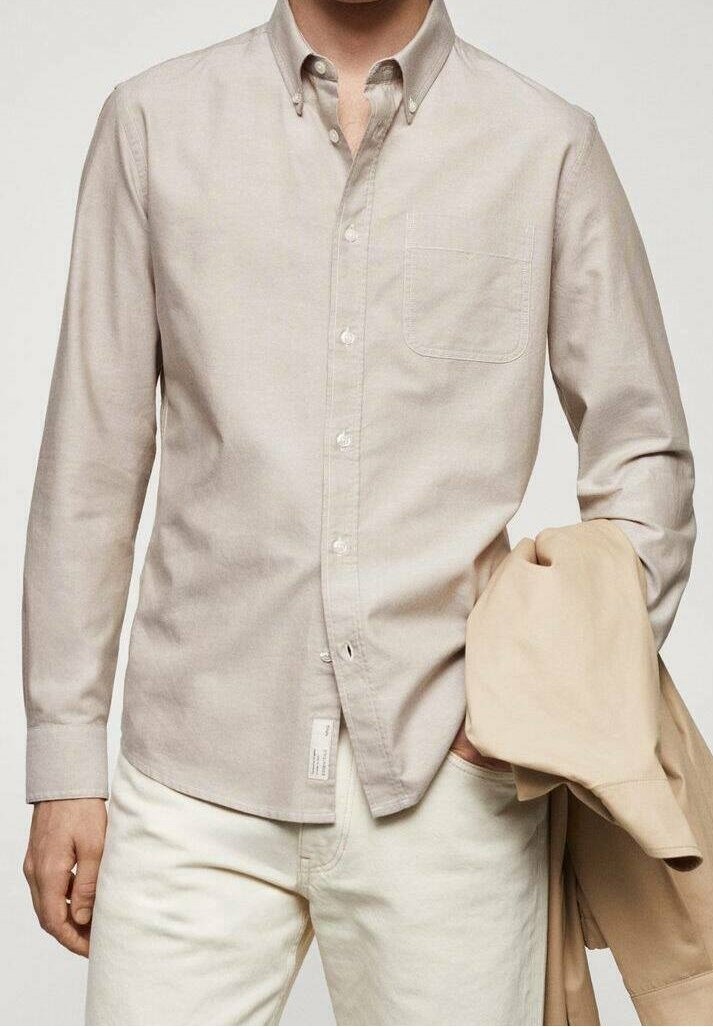 Homme portant une chemise beige boutonnée avec une poche poitrine, tenant une veste marron clair dans la main gauche et portant un pantalon blanc cassé.