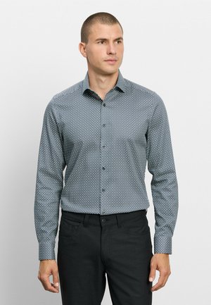 Mann trägt ein eng anliegendes blau gemustertes Langarmhemd und schwarze Hose, schaut leicht zur Seite.