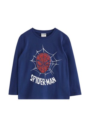 Smilende dreng står med hænderne i lommerne iført en blå Spider-Man langærmet skjorte, cowboybukser og blå sneakers mod en ensfarvet baggrund.