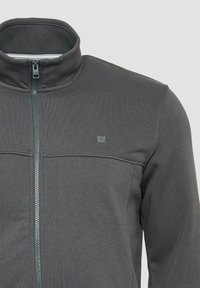 Giacca a zip grigia scura con colletto alto, realizzata in tessuto liscio. Presenta un logo sottile sul petto e cuciture a contrasto.