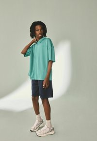 Turkos kortärmad skjorta med krage och en texturerad meshdesign, matchad med marinblå shorts och vita ankelsockor i kombination med ljusa sneakers.