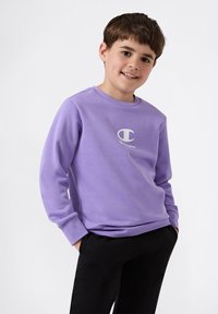 Paarse sweater met een ronde hals, lange mouwen en een wit logo op de borst. Gecombineerd met zwarte broek, met een casual uitstraling.
