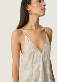 Woman with long dark hair wearing silver hoop earrings and a silver floral-patterned sleeveless V-neck top, facing right.