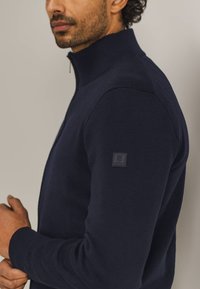 Dunkelblauer Zip-Hals-Pullover aus strukturiertem Stoff mit taillierter Passform. Verfügt über ein Logo-Patch auf der linken Schulter.