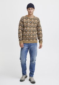 Gebreide trui in bruin met een ruitpatroon in bruin, zwart en wit, gecombineerd met blauwe jeans en grijze schoenen. Model draagt een donkere beanie.