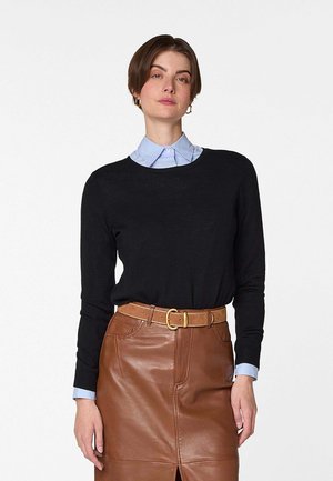 Frau trägt einen schwarzen Langarmpullover über einem hellblauen Hemd mit Kragen, kombiniert mit einem braunen Lederminirock und Gürtel, steht vor weißem Hintergrund.