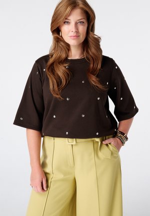 Femme aux longs cheveux ondulés porte un haut noir à manches courtes clouté et un pantalon taille haute jaune clair, debout avec une main dans la poche.
