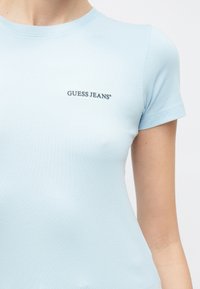 Svetlo modra oprijeta majica iz mehkega blaga, z okrolim izrezom in kratkimi rokavi, na sprednji strani z temno modrim napisom "GUESS JEANS".