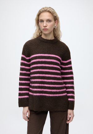 Jeune femme aux cheveux blonds porte un pull marron avec des rayures horizontales roses et un pantalon marron assorti sur fond uni.