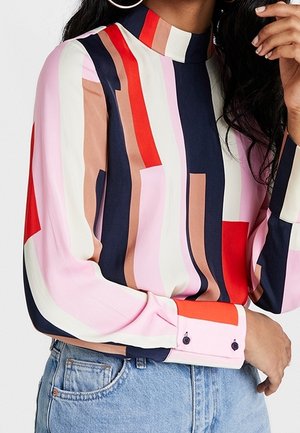 Kvinde iført en flerfarvet, stribet bluse med lange ærmer og høj krave samt lyseblå jeans, der holder hånden nær sit ansigt.