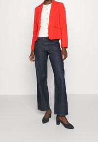 Blazer rouge, chemise blanche à boutons, jean large foncé. Tissu lisse, design structuré, coupe ajustée. Chaussures à talons hauts bleues.
