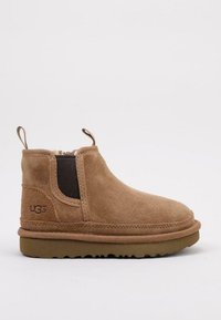 Botín de ante color marrón con panel lateral elástico, punta redondeada y suela de goma. Cuenta con una lengüeta en el talón y el logo de UGG a un lado.