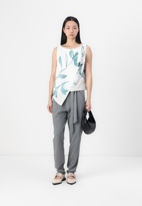 Haut sans manches blanc avec imprimé de feuilles vertes, design asymétrique ; associé à un pantalon taille haute gris, des ballerines beiges et un sac noir.