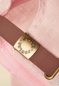 Primo piano di un cappello in tessuto rosa con una cinghia regolabile marrone dotata di una fibbia metallica dorata incisa con la scritta "JOULES".