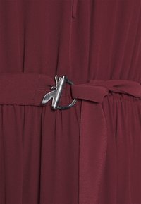 Robe bordeaux avec une texture plissée, dotée d'une boucle en métal argenté et d'une ceinture en tissu assortie pour cintrer la taille.