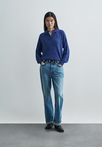 Blauer gestrickter Pullover mit Kragen, kombiniert mit hellblauen Jeans in lockerer Passform. Das Outfit wird durch einen schwarzen Gürtel und schwarze Schuhe mit Absatz ergänzt.